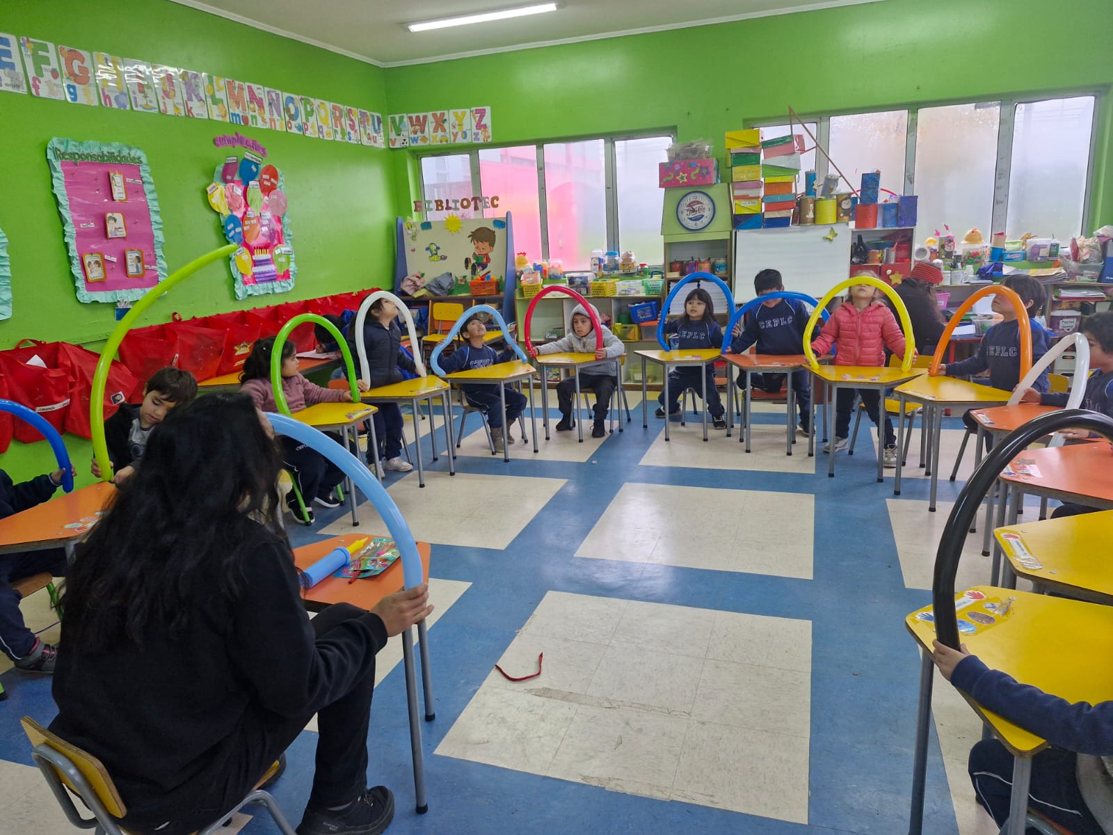 Niños sentados en sus sillas con sus mesas, formando un cirulo. Cada uno iene un arco hecho con globos de los largos