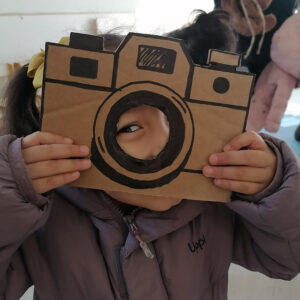 Niñas sosteniendo una cámara fotográfica dibujada en un cartón café. El circulo central de la cámara se encuentra recortada y ella esta mirando con un ojos por el orificios.