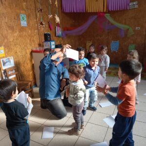 Niños de pie jugando con hojas blancas de papel, mientras un profesor esta arrodillado en el suelo con una hoja blanca.