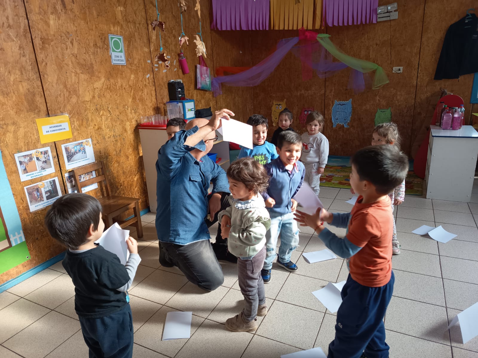 Niños de pie jugando con hojas blancas de papel, mientras un profesor esta arrodillado en el suelo con una hoja blanca.