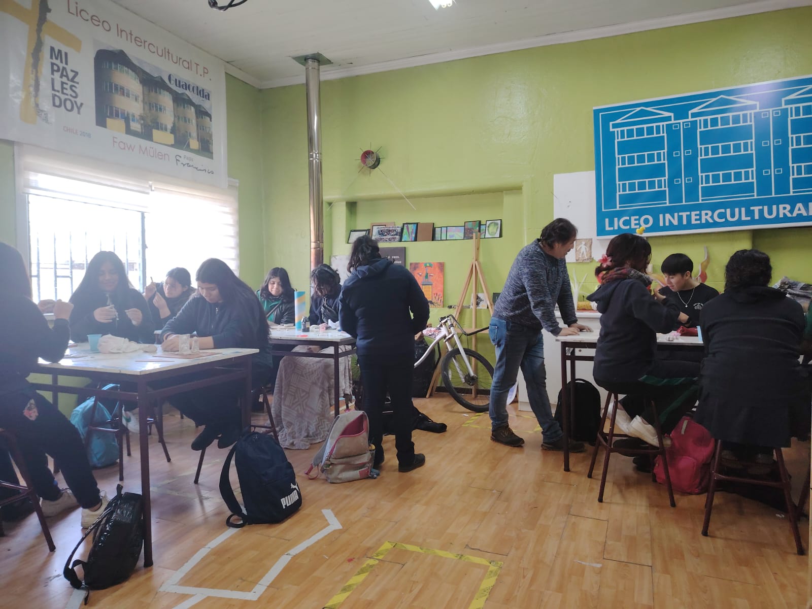 Jovenes sentados en mesas de trabajo mientras hacen manualidades y un profesor los supervisa