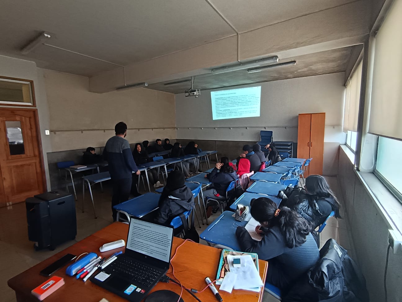 Jovenes sentados en mesas azules, mientras un profesor proyecta una presentación