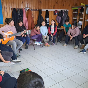 Niños sentados en sillas formando un círculo, mientras profesora toca la guitarra