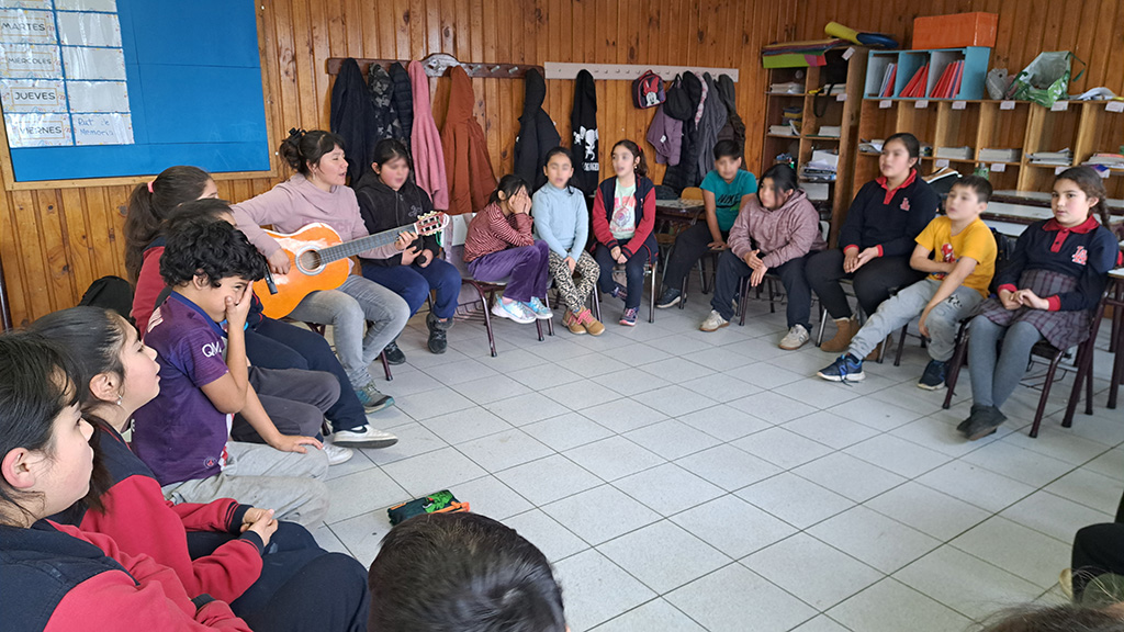 Niños sentados en sillas formando un círculo, mientras profesora toca la guitarra