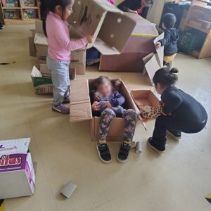 Niños jugando con cajas de cartón. Una niña se encuentra dentro de una caja con una muñeca