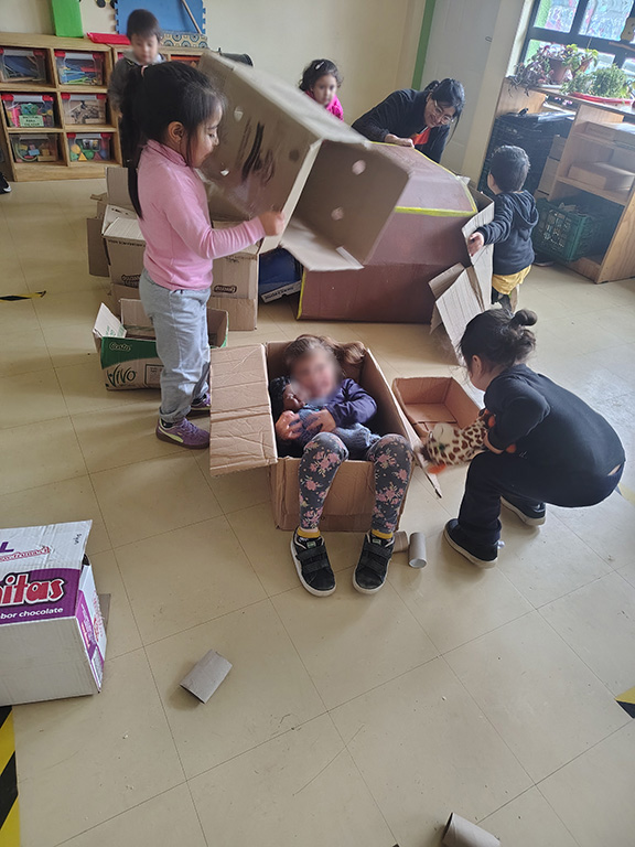 Niños jugando con cajas de cartón. Una niña se encuentra dentro de una caja con una muñeca
