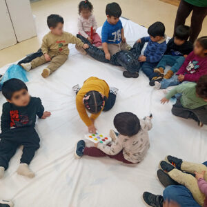 Niños sentados en el suelo sobre una tela blanca. Algunos están jugando