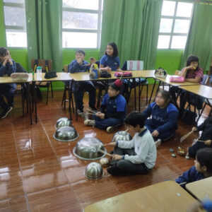 Niños sentados en mesas y suelo. Los que están en el suelo están tocando bowls metálicos con baquetas de madera