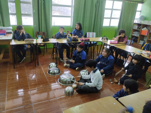 Niños sentados en mesas y suelo. Los que están en el suelo están tocando bowls metálicos con baquetas de madera
